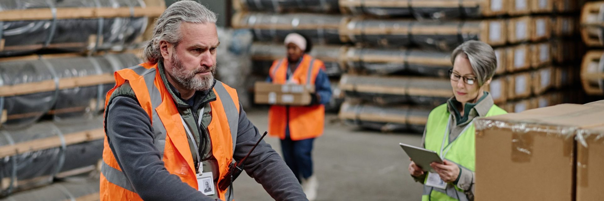 Mature worker in protective workwear loading cargo with loader in storage room with foreman using tablet pc in background